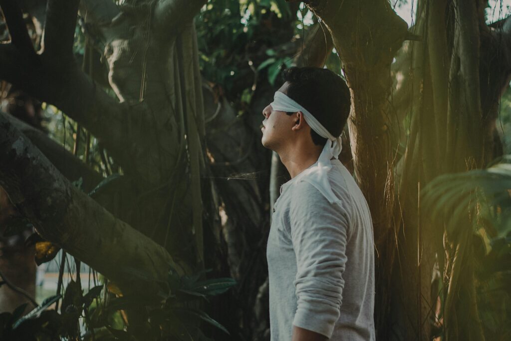 A blindfolded Asian man dressed in white stands under a large tree with many branches.
