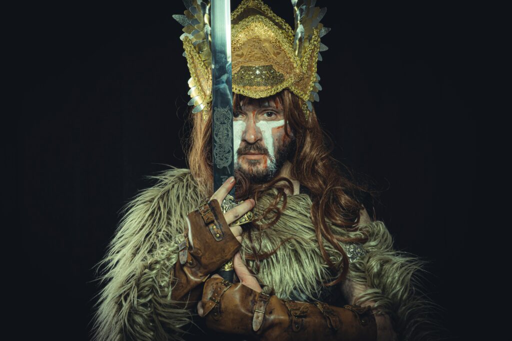 A white man with face paint and an elaborate headdress holds up a sword with a solemn expression.