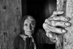 An old woman, dressed in black, stands with her hand resting on a wooden doorframe.