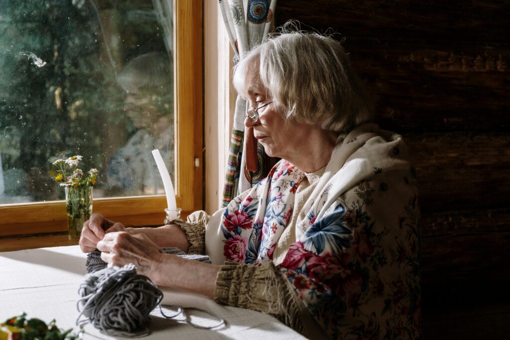 An old woman with glasses sits at a table, with a ball of yarn to her side.