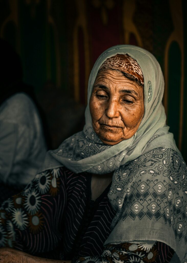 An old woman dressed in silvery blue, with her head covered, gazes downwards.
