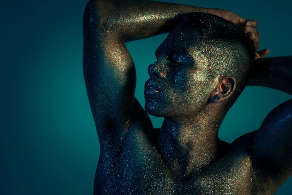 A young man covered in glitter and shining with blue light poses for the camera.